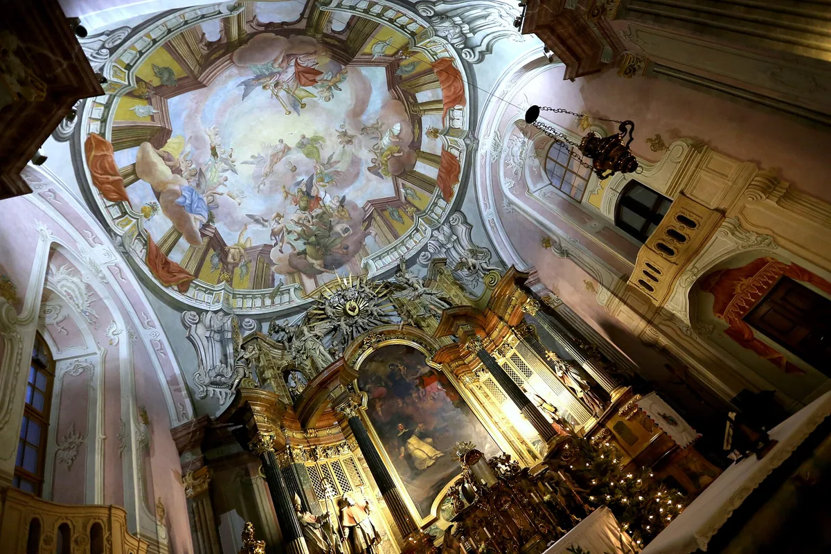 A gilded Baroque high altar with a painting above it and a ceiling fresco of figures among clouds, seen from below