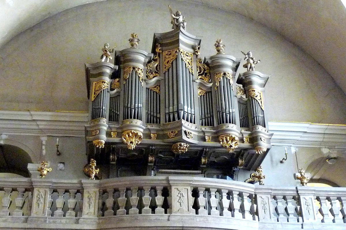 The silver pipes of a church organ in a gilded case on a stone balcony, beneath a pale vaulted ceiling