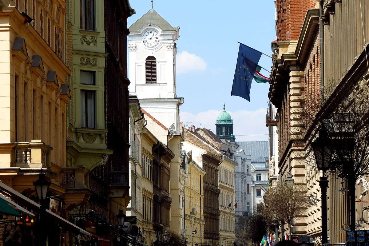 Váci utca in Budapest, a narrow pedestrian street between tall nineteenth-century buildings, with the white clock tower of the church rising at the end