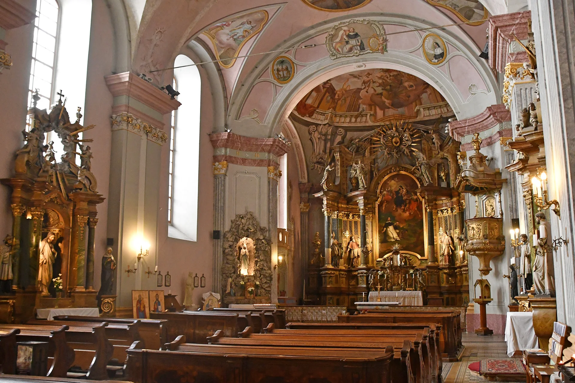 The nave of a Baroque church in Budapest, with rows of dark wooden pews facing a gilded high altar, pink-painted vaults with frescoes and tall arched windows